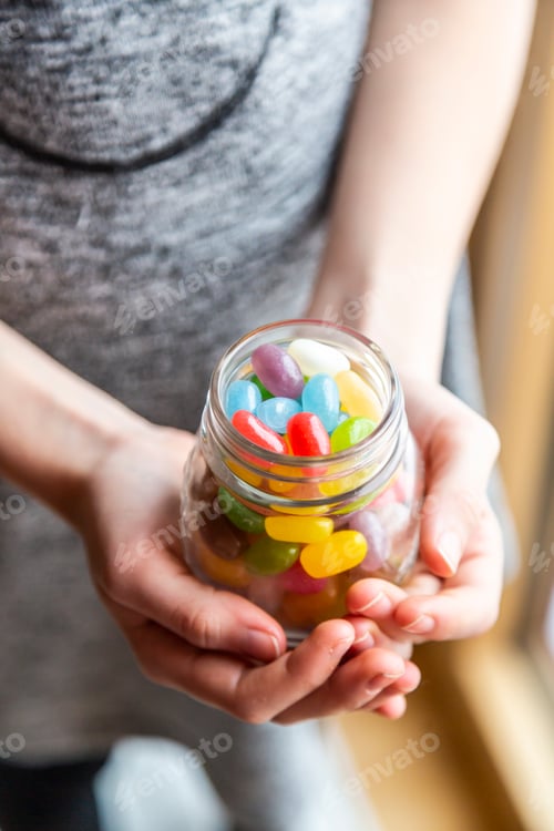 Preview: Glass of colourful sweet jellybeans on white wood, hands holding glass