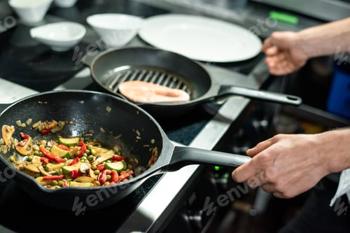 Preview: Hands of male chef roasting piece of salmon and vegetable stew on frying pans