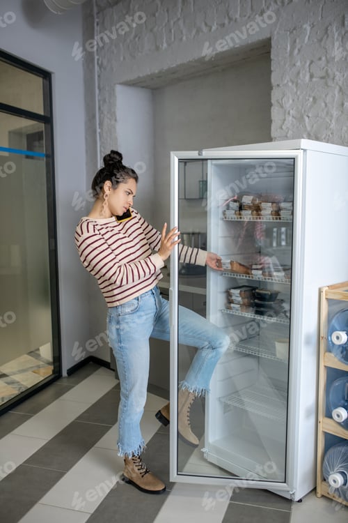 Preview: Young woman taking food container from fridge, talking on mobile