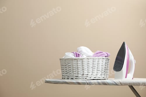 Vista previa: New modern iron and basket with laundry on board against beige background, space for text
