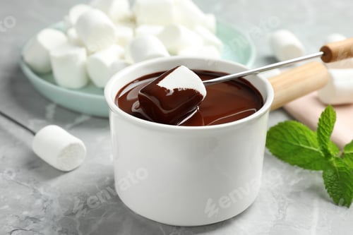 Preview: Dipping marshmallow into fondue pot with dark chocolate on marble table, closeup