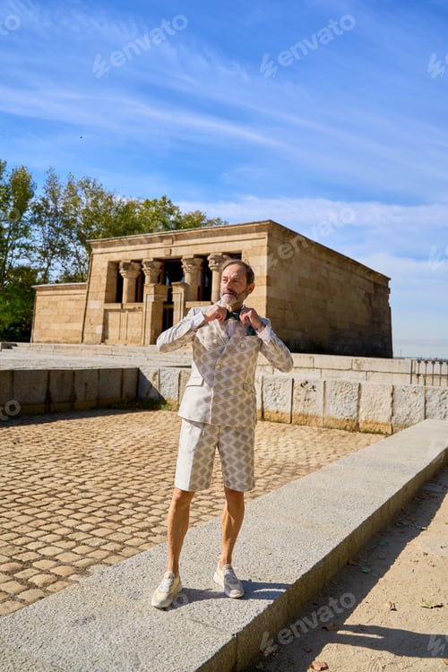 Preview: Man in patterned suit adjusting bow tie near ancient stone temple outdoors