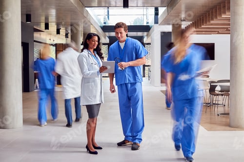 Preview: Male And Female Doctors In White Coat And Scrubs Looking At Digital Tablet n Busy Hospital