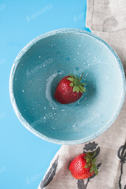 Preview: One red juicy strawberry in a blue deep plate on a blue background. Next is a linen wipes. Top view.