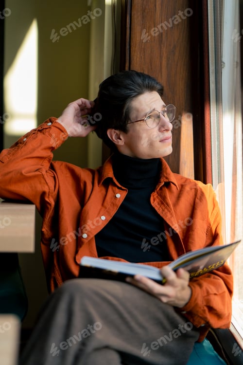 Preview: a young guy in glasses sits in a cafe with a book and looks thoughtfully out window