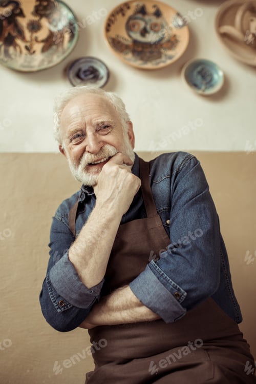 Preview: Senior potter in apron thinking and standing against wall with hanging pottery goods