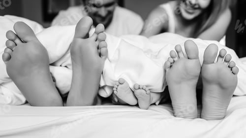 Preview: black and white picture of legs of family with infant son under blanket in bed at home