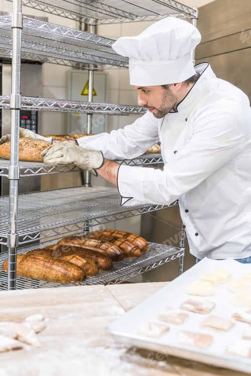 Preview: adult male baker in white chefs uniform putting fresh hot bread at rack