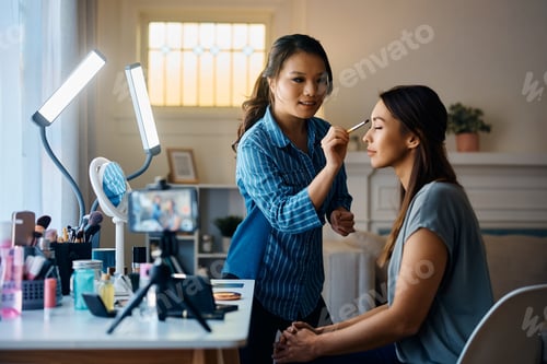 Preview: Professional Asian beautician applying makeup on young woman's face.