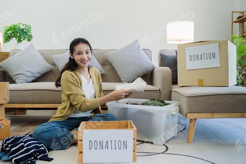 Preview: Asian young woman packing clothes at home, putting on stuff into donate box with second hand clothes