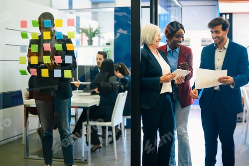 Preview: Group of positive multiracial colleagues discussing documents while standing against glass wall
