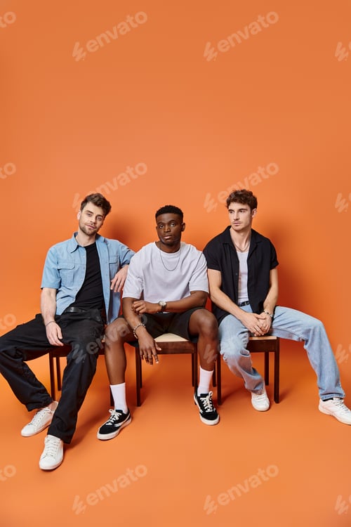 Preview: good looking multicultural male models in casual urban attires sitting on chairs on orange backdrop