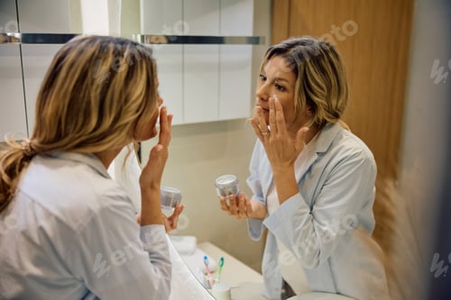 Preview: Reflection in a mirror of a woman applying face cream in the morning.