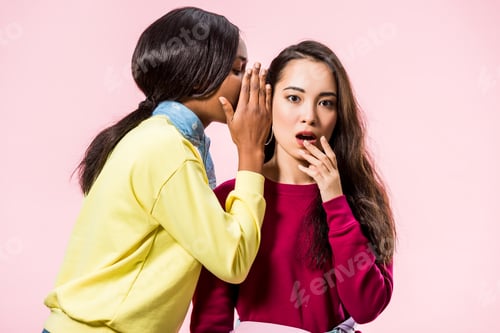 Preview: african american woman telling secret to her shocked asian friend isolated on pink