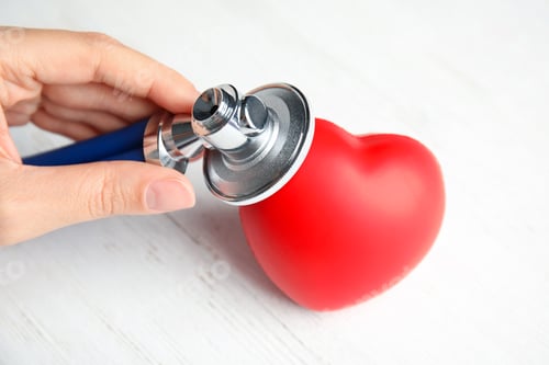 Preview: Woman holding stethoscope near red heart on wooden table, closeup. Cardiology concept