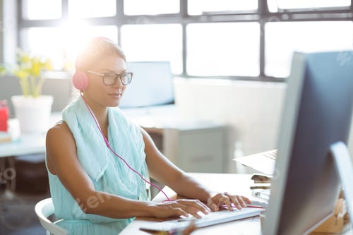 Preview: Woman typing on keyboard at office desk, wearing pink headphones and glasses, copy space