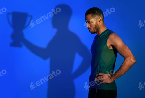 Preview: Young black sportsman dreaming about winning trophy, shadow of champion holding prize on blue studio