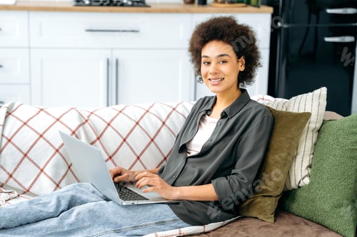 Preview: Satisfied african american woman with short curly hair, in stylish wear, sit on a sofa in living
