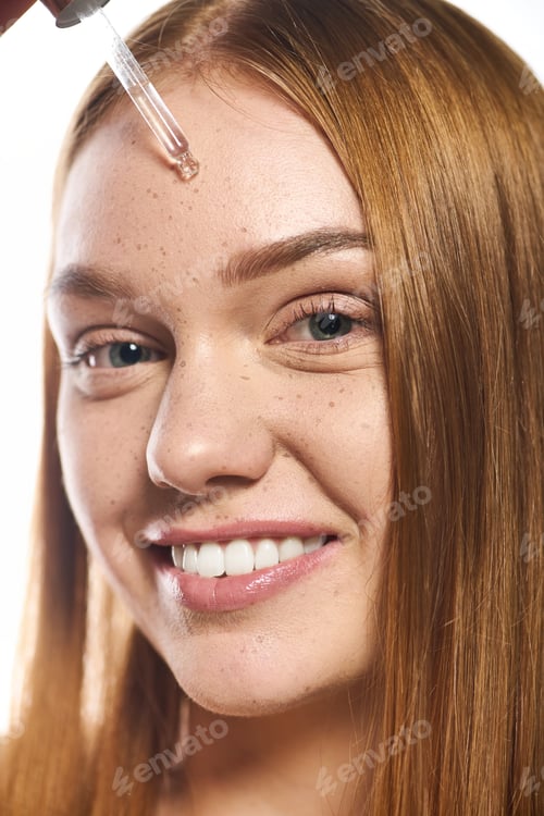 Preview: Young woman applying skincare product, smiling with radiant skin in a bright setting