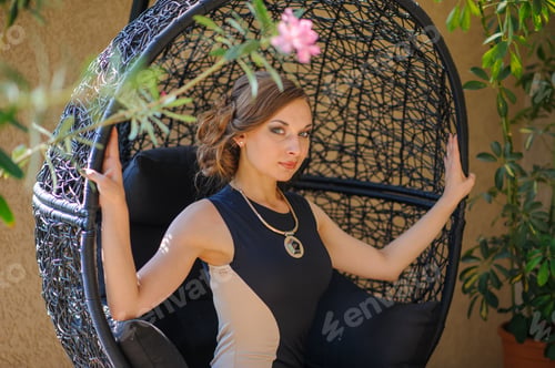 Preview: Young beautiful girl in a dress sits on a swing chair on the terrace.