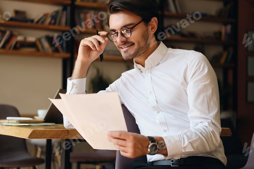 Preview: Young smiling businessman in eyeglasses and white shirt happily working with papers in modern office
