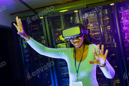 Preview: Happy african american female computer technician wearing vr headset and working in server room