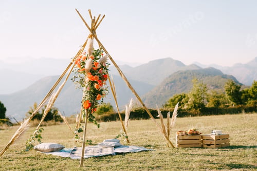 Preview: Boho-style wedding arch stands near a bedspread with pillows and boxes of food in a clearing