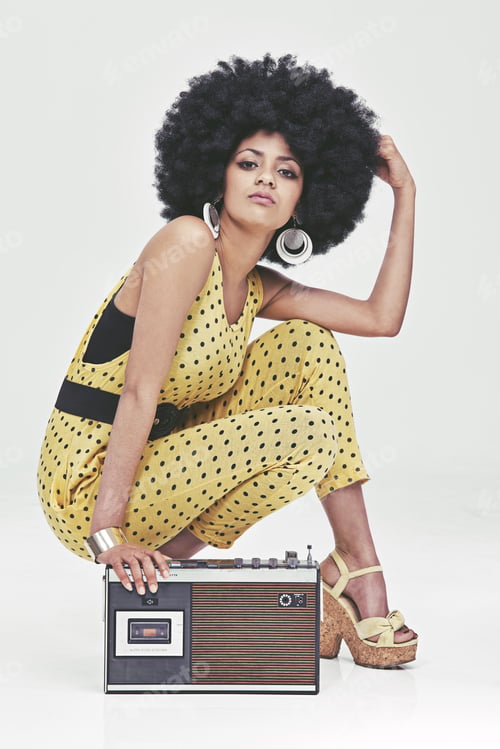 Preview: A young woman wearing a 70s retro jumpsuit sitting by a cassette player striking a pose