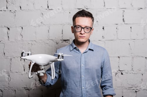 Preview: Young stylish Man in glasses holding the quadcopter drone DJI Phantom 4 on a grey brick wall