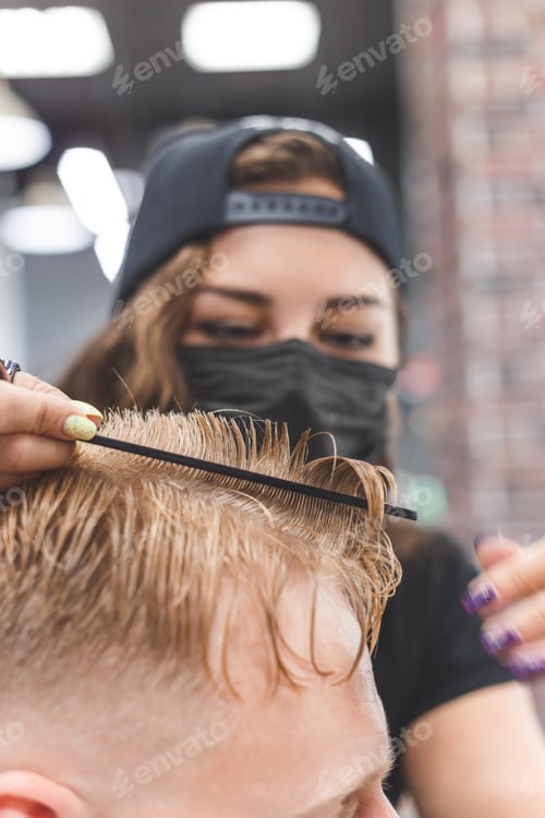 Preview: Woman barber cutting hair to a bearded man in face mask. Quarantine haircut concept.