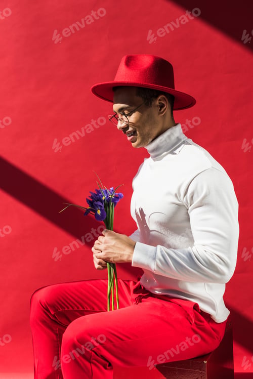Preview: stylish mixed race man in hat and glasses holding flowers and smiling on red
