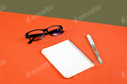 Preview: Blank white notebook, eyeglasses and pen on orange and green background.
