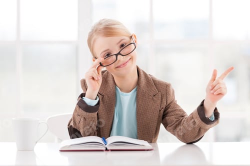 Preview: Look at that! Cheerful little girl in glasses and formalwear sitting at the table and pointing away