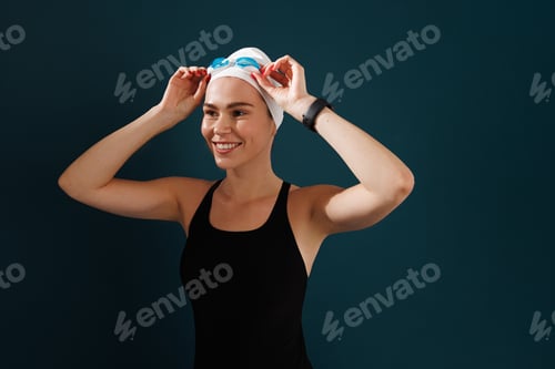 Preview: Smiling woman in swimwear adjusting her goggles while standing at blue backdrop
