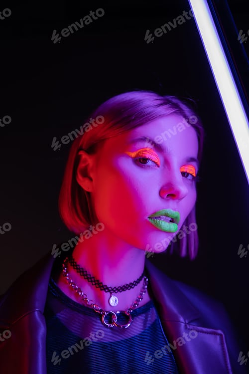 Preview: trendy woman with bright neon makeup looking at camera near purple fluorescent lamp on black