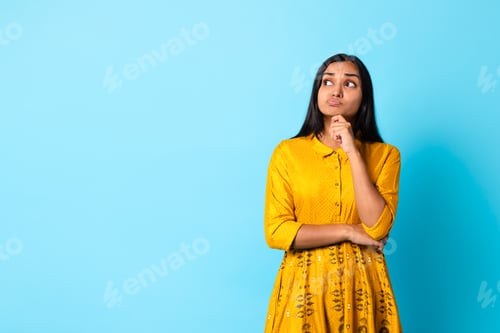 Preview: Thoughtful young Indian lady in traditional yellow dress, blue backdrop