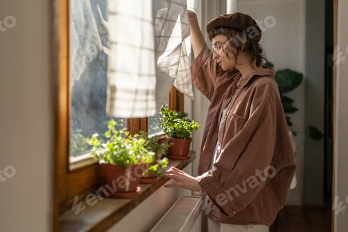 Preview: Interested teenage girl looks out window with green plants in pots, waiting visit by friend at home.