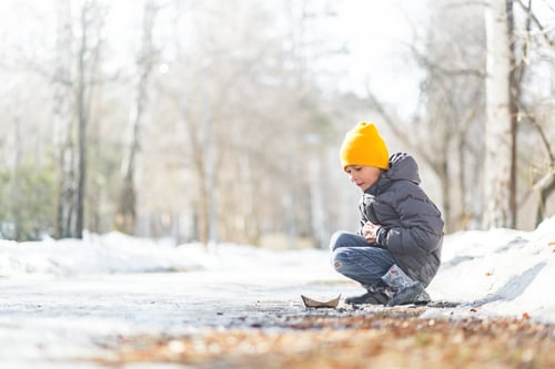 Preview: Boy in grey jacket and yellow knitted hat playing with paper boat near puddle in spring forest
