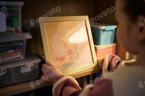 Preview: Young Caucasian Girl Holding Framed Drawing of Family in Cozy Room