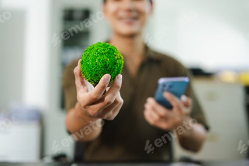 Preview: Young Asian man holding a globe and smartphone at desk, symbolizing green technology,