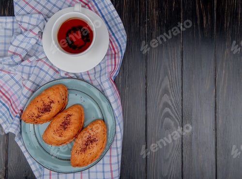 Preview: top view of badamburas in plate and cup of tea on saucer on plaid cloth and wooden background with