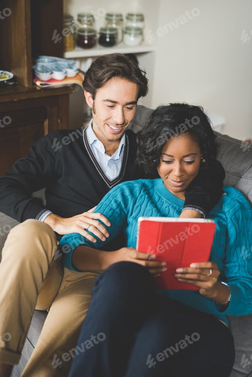 Preview: Young handsome mixed race couple sitting on the couch smiling and having fun using a tablet