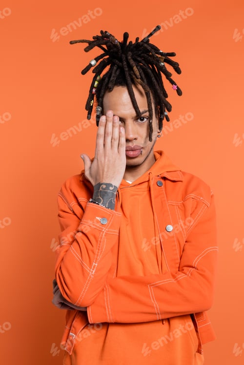 Preview: tattooed and multiracial man with dreadlocks covering half of face isolated on coral