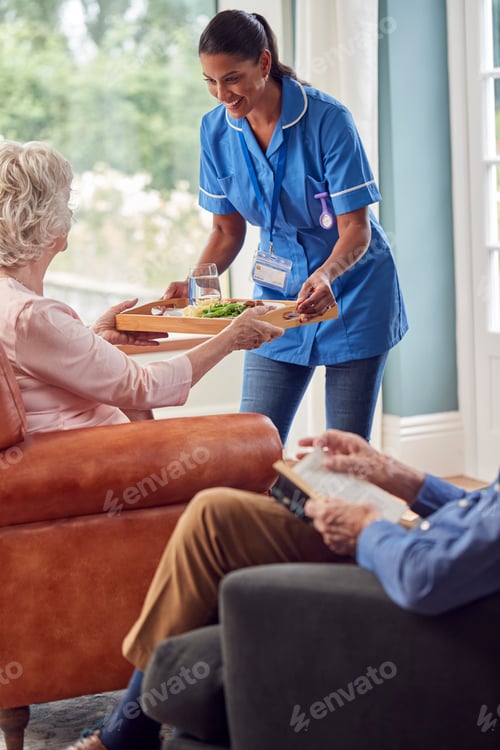 Preview: Female Care Worker In Uniform Bringing Meal On Tray To Senior Woman Sitting In Lounge At Home