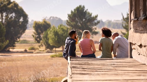 Preview: Rear View Of Group Of Friends On Vacation Sitting On Porch Of Countryside Cabin Drinking Beer