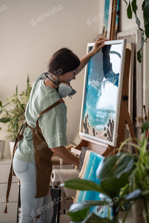 Preview: Focused artistic woman putting canvas with epoxy resin painting in liquid art technique on easel.