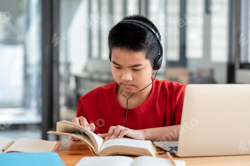 Preview: Asian boy wearing headphones while reading a book the laptop placed on the table in the room.