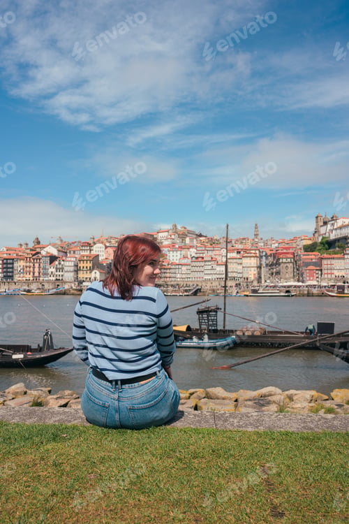 Preview: Young woman sitting on riverside in Porto, Portugal overlooking traditional boats and cityscape