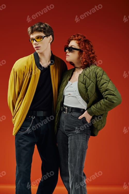 Preview: beautiful fashionable couple with chic sunglasses in cozy attires posing on red backdrop together