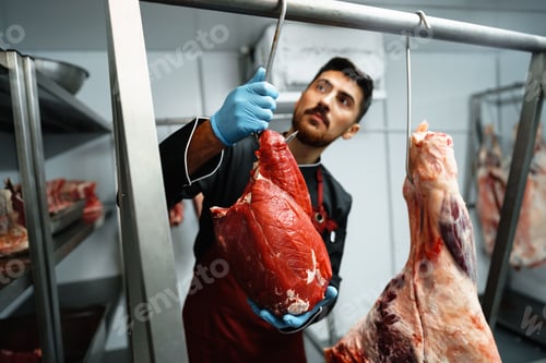 Preview: Male butcher standing by meat hanging on hook in meat shop
